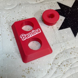 May include: A red plastic spool holder with two holes and the word "Bernina" printed on it. There is a smaller red plastic spool holder next to it.