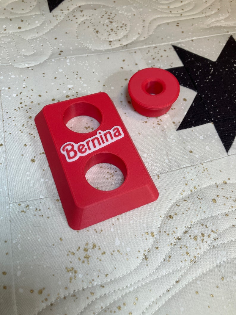 May include: A red plastic spool holder with two holes and the word "Bernina" printed on it. There is a smaller red plastic spool holder next to it.