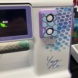 May include: A white and purple sewing machine with a blue and white honeycomb pattern on the side. The machine has a digital display with the word "eco" in green. The machine also has a button with two circles on it. The machine has the text "Yaya Han Edition" and "bernette SWISS DESIGN" on it.