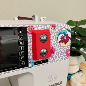 Może przedstawiać: Zbliżenie na białą maszynę do szycia z czerwonym panelem sterowania. Maszyna ma wyświetlacz cyfrowy i wiele przycisków. Tekst na maszynie brzmi "(KAffe) SWISS ENGINEERING BERNINA KAFFE EDITION B 770 QE PLUS".