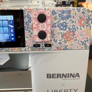 Może przedstawiać: Biała maszyna do szycia Bernina z kwiatowym wzorem Liberty w odcieniach różu, błękitu i żółci. Maszyna posiada cyfrowy ekran, różowy prostokątny panel sterowania z dwoma czarnymi pokrętłami oraz napis Bernina Swiss Engineering x Liberty.