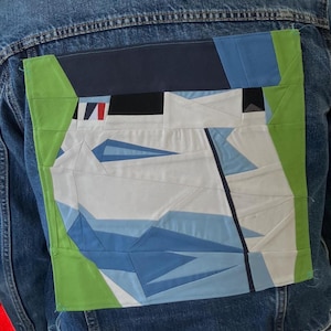May include: A denim jacket with a large, geometric patch on the back. The patch features a design with white, blue, black, and green shapes, creating an abstract pattern. The patch is bordered with green fabric.