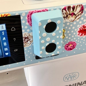 May include: A light blue sewing machine with a white polka dot design and floral accents. The machine has a digital display with a question mark, a clear button, and a heart icon. The machine also has a blue and white polka dot accessory holder.