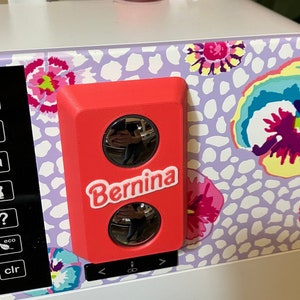 May include: A red sewing machine accessory with two round, reflective windows and the word "Bernina" printed in white.
