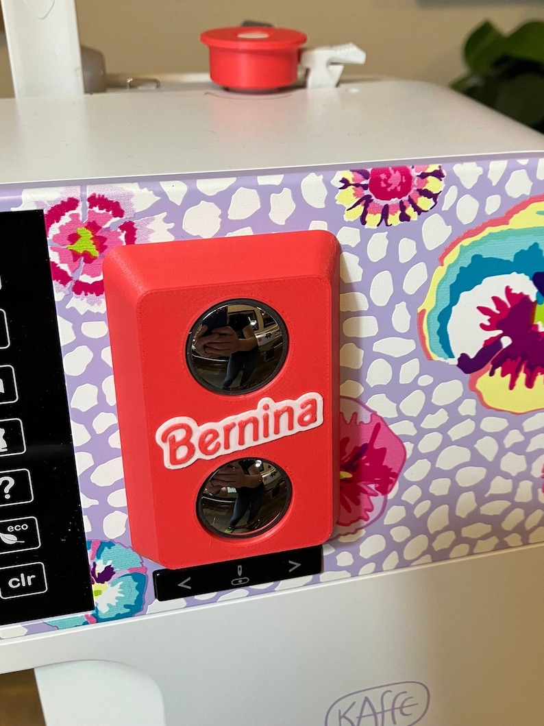 May include: A red sewing machine accessory with two round, reflective windows and the word "Bernina" printed in white.
