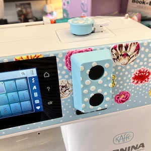 Könnte beinhalten: Eine weiße Nähmaschine mit einem blauen und weißen Blumenmuster. Die Maschine hat ein digitales Display mit Stichoptionen und einen blauen Knopf. Die Maschine hat auch einen blauen Knopf mit zwei Löchern.