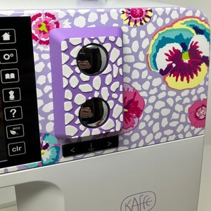 Peut inclure: Gros plan d'une machine à coudre violette avec un motif floral. La machine possède un écran numérique avec des boutons pour différentes fonctions. La machine possède également un bouton violet avec deux fenêtres rondes et transparentes.