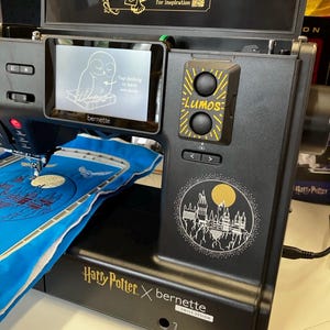 Pode incluir: Máquina de costura preta com um ecrã que exibe um gráfico de coruja. A máquina tem a marca "LUMOS" e "Harry Potter x bernette". Um gráfico de coruja dourada e o texto "Scan QR code for inspiration" também são visíveis.