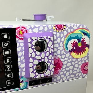 Puede incluir: Primer plano de una máquina de coser de color morado con un diseño floral. La máquina tiene una pantalla digital y un panel de control con botones para diversas funciones. El panel de control tiene una cubierta morada con una superficie texturizada y dos botones redondos.