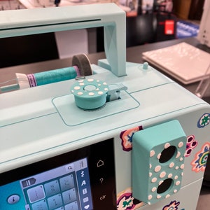 May include: A light blue sewing machine with a digital display and a variety of stitch options. The machine has a white polka dot design and the text "KAFFE FINA" is visible on the front.
