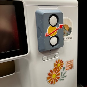 May include: A white sewing machine featuring a grey 3D-printed attachment with two white circular buttons and a yellow planet graphic with a red ring. Two stickers are applied to the machine: one reading "CASHMERE COMMUNITY FARMERS MARKET" with orange flowers, and another reading "EY CACTUS BOUTIQUE" with a desert landscape.