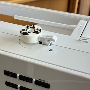 May include: White sewing machine with a paw print knob and a silver hook. The machine has a digital display with buttons labelled "mem 0" and "mem 1".