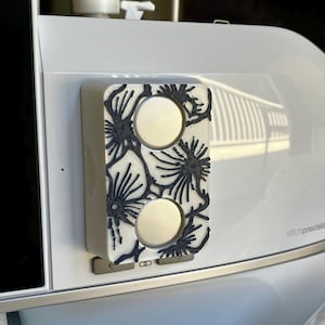 Może przedstawiać: Prostokątny, kremowy obiekt z dwoma okrągłymi otworami i czarnym kwiatowym wzorem. Obiekt jest przymocowany do białego urządzenia. Kwiatowy wzór jest skomplikowany i szczegółowy.