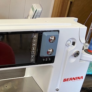 May include: A white Bernina sewing machine with a blue and silver control panel and a red logo. The machine has a digital display and a needle threader.
