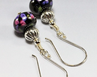 May include: A pair of dangle earrings with black glass beads featuring a floral pattern in shades of purple and pink. The beads are attached to silver-toned metal posts with gold-toned accents.