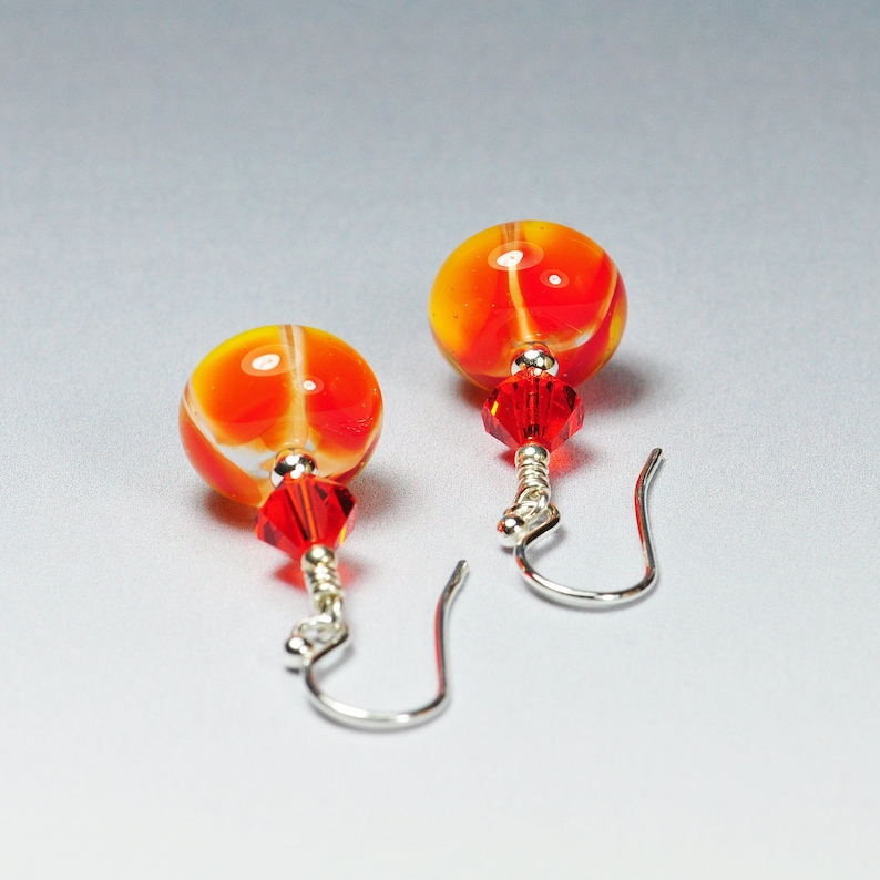 May include: A pair of handmade earrings featuring vibrant orange and yellow glass beads with red accents. Each earring has a red crystal bead and silver-colored ear wires. The earrings are designed for pierced ears.