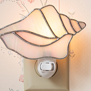 May include: A white seashell-shaped nightlight with a silver metal frame. The nightlight is plugged into an electrical outlet.