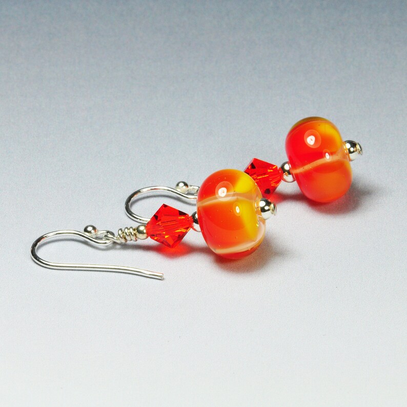 May include: A pair of dangle earrings featuring orange and yellow glass beads with a white stripe. The earrings also have red crystal accents and silver-colored ear wires. The beads have a glossy finish.