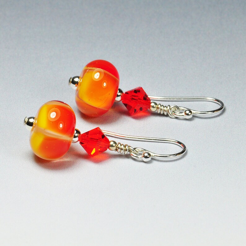May include: A pair of handmade earrings featuring orange and yellow glass beads with a silver accent. The earrings also have a red crystal and silver ear wires. The earrings are a vibrant accessory.