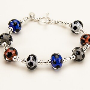 May include: A beaded bracelet featuring colorful glass beads with black polka dots. The beads are in shades of blue, gray, and copper, strung on a silver chain with a toggle clasp. A stylish accessory.