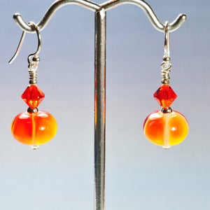 May include: A pair of handmade earrings featuring orange and yellow glass beads with a silver hook. Each earring has a faceted red crystal bead above the glass bead. The earrings are displayed on a silver earring stand.
