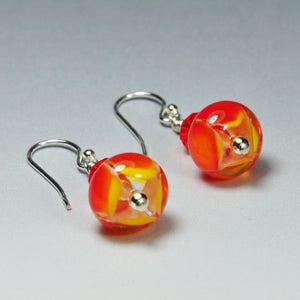 May include: A pair of handmade earrings featuring vibrant orange and yellow glass beads with silver accents. The earrings have a hook design and a small red bead at the top. The glass beads have a spherical shape.