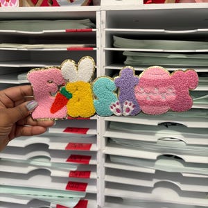 May include: A colorful Easter-themed felt decoration spelling out "2023". The design features a pink bunny, a carrot, a yellow bunny, a blue bunny, a purple bunny, and a pink egg. The edges are trimmed with gold thread.