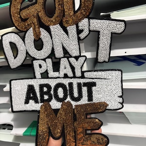 May include: A decorative patch with the words "GOD DON'T PLAY ABOUT ME" in gold and white sequins. The patch is black around the edges. The patch is held up in front of a white background.