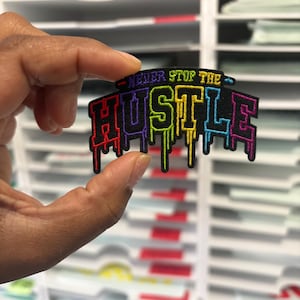 Può includere: Una toppa colorata con la scritta "NEVER STOP THE HUSTLE" in un font a goccia. La toppa presenta uno sfondo nero con testo dai colori arcobaleno. La toppa è tenuta davanti a uno sfondo bianco.