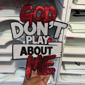 May include: A decorative sign with the text "GOD DON'T PLAY ABOUT ME". The word "GOD" and "ME" are in red sequins, while "DON'T PLAY ABOUT" is in black and white sequins. The sign is held in front of a white shelf.