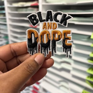 Puede incluir: Parche bordado con las palabras "BLACK AND DOPE" en letras negras y doradas. La palabra "DOPE" tiene un efecto de goteo. El parche se sostiene en una mano sobre un fondo de estanterías blancas.