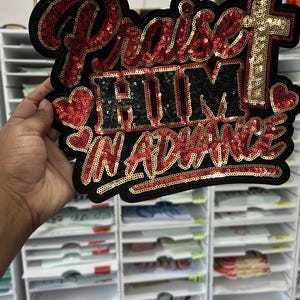 May include: A large decorative patch featuring the text Praise Him In Advance in red and black sequins. The design includes two red sequined hearts and a gold sequined cross on the right side, all outlined in black with a thick black border.