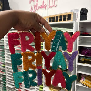 May include: Colorful, textured fabric letters spelling "FRIYAY" in various colors, including red, orange, teal, and purple. Some letters feature ruler and pencil designs. The letters are trimmed with gold fabric. The sign "Design Sisters & Blanks LLC" is visible in the background.