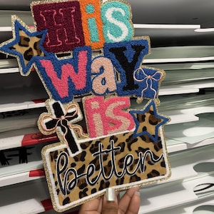 May include: A decorative patch with the words "His Way Is Better" in various colours and fonts. The patch features a leopard print background, gold glitter trim, and star and cross accents. The patch is held by a hand.