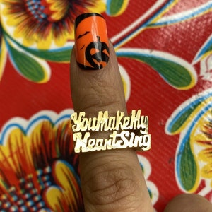Puede incluir: Anillo de oro con las palabras "You Make My Heart Sing" grabadas. El anillo se lleva en un dedo con esmalte de uñas de temática de Halloween.