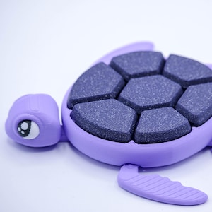 May include: A purple turtle-shaped object with a segmented, dark gray shell. The turtle has a detailed head with an eye and flippers. The shell is composed of several hexagonal pieces. The object is likely a decorative item or a toy.