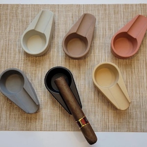 Puede incluir: Seis reposacigarros de cerámica en varios colores, incluyendo gris, beige, marrón y negro. Un reposacigarros tiene un cigarro apoyado en él.