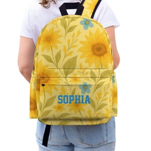 May include: Yellow backpack with a floral sunflower pattern and the name "SOPHIA" in blue block letters. The backpack has a front pocket, black straps, and a black top handle. The background is white.