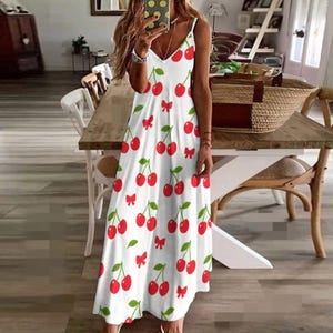May include: White sundress with a red cherry pattern. The dress features a V-neckline and spaghetti straps. Red bows and green leaves are interspersed among the cherries. The dress is a midi length.