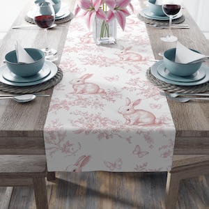 May include: A white table runner featuring a pink floral design and pink bunnies. The runner is adorned with butterflies and has a delicate, vintage style.
