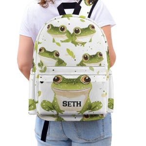 Puede incluir: Mochila blanca con un patrón repetido de ranas verdes y hojas. El bolsillo delantero muestra una gran rana con el nombre "SETH" en texto negro. La mochila tiene correas negras y un asa negra.