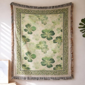 May include: A rectangular woven throw blanket with a repeating pattern of green four-leaf clovers on a cream background. The border has a green Celtic knot design. The edges are fringed with brown and grey threads.
