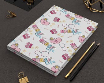 May include: A white notebook with a pink and blue pattern featuring the text "Nurse Life" repeated throughout. The pattern also includes hearts, flowers, stethoscopes, and syringes.