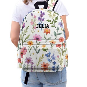 Puede incluir: Mochila blanca con un estampado floral colorido con flores rosas, naranjas, moradas y azules. El nombre "JULIA" está impreso en negro. La mochila tiene correas negras y un bolsillo frontal.