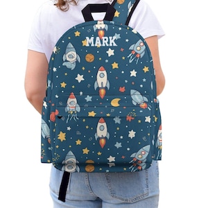 Puede incluir: Una mochila azul marino con un diseño espacial de cohetes, estrellas y planetas. El nombre "MARK" está impreso en blanco en la parte superior. La mochila tiene un asa y correas negras.