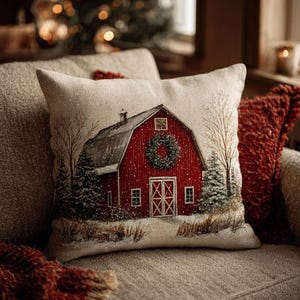 May include: A square cushion with a festive red barn design, complete with a green wreath, set against a snowy winter landscape. The cushion is part of a seasonal collection, ideal for adding a touch of warmth to a living space.