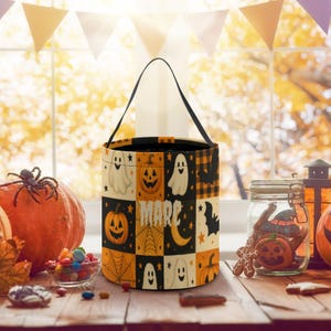 Könnte beinhalten: Ein Halloween-Eimer mit schwarzem Griff. Der Eimer hat ein Patchwork-Design mit Kürbissen, Geistern, Fledermäusen und dem Wort "MARC". Das Farbschema umfasst Orange, Schwarz und Weiß.