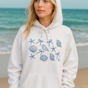 May include: White hooded sweatshirt with a drawstring and a front pocket. The sweatshirt features a blue seashell and starfish pattern. The model is standing on a beach with the ocean in the background.