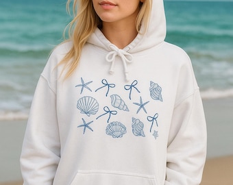 May include: White hooded sweatshirt with a drawstring and a front pocket. The sweatshirt features a blue seashell and starfish pattern. The model is standing on a beach with the ocean in the background.