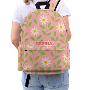 May include: Pink backpack featuring a daisy and leaf design. The backpack has a front pocket with the name "SOPHIA" printed on it. It has black straps and a black handle.